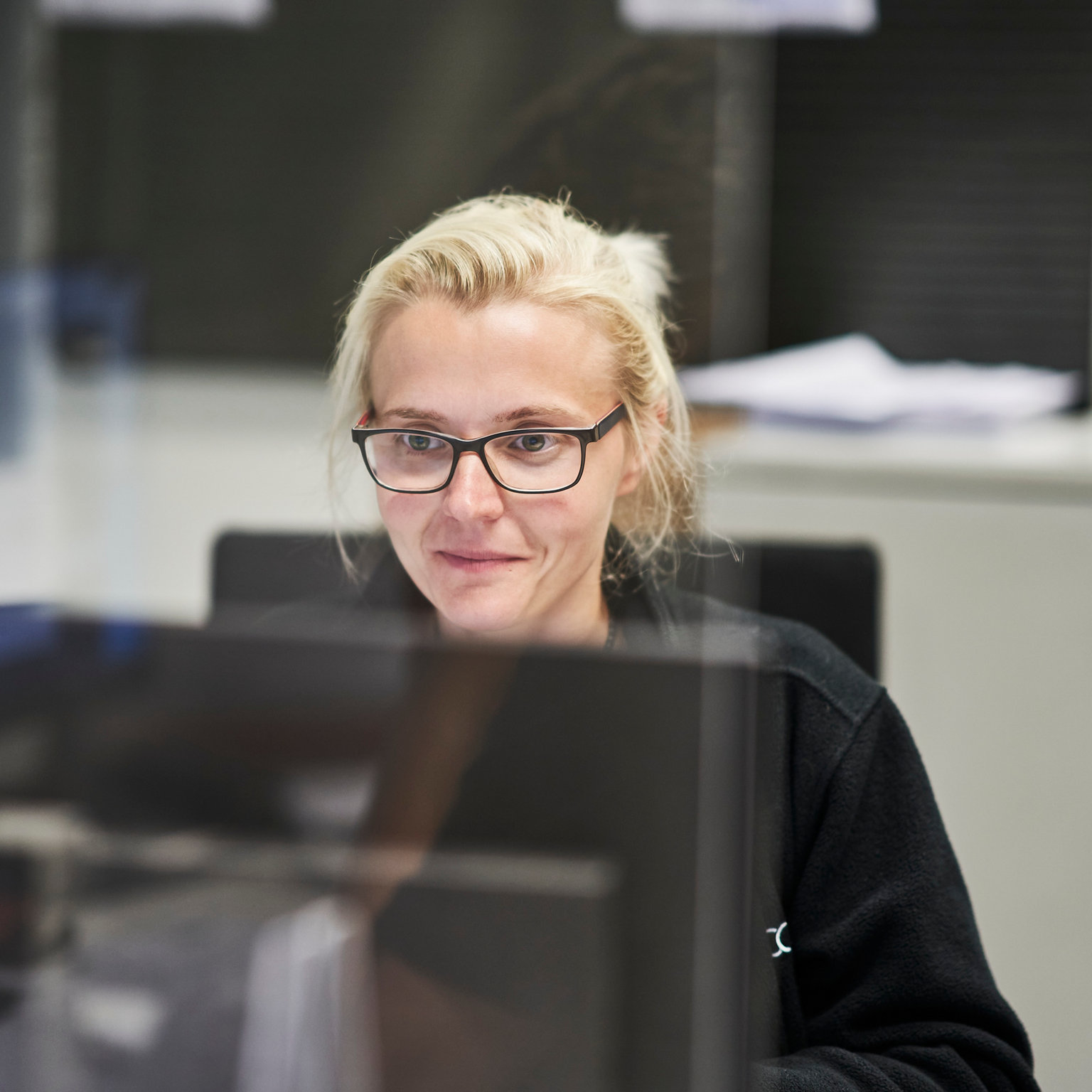 Blonde Mitarbeiterin mit Brille sitzt in einem Büro und schaut auf den Computer-Bildschirm
